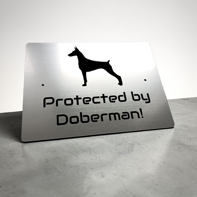Stříbrná tabulka se siluetou dobrmana a anglickým nápisem Protected by Doberman