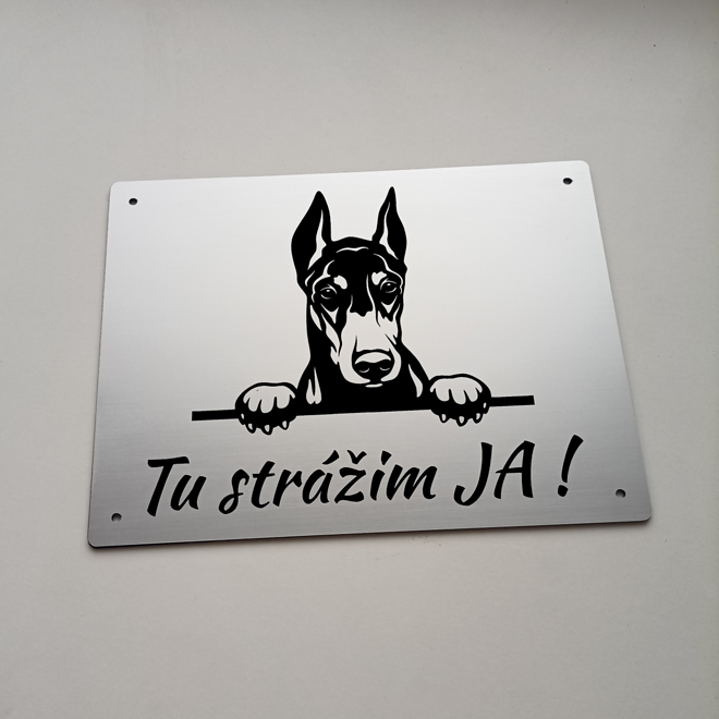 Stříbrná tabulka s vykukujícím dobrmanem a nápisem Tady hlídám já